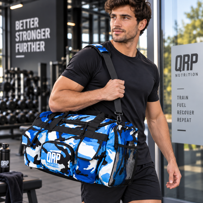 A man in a black t-shirt and shorts carrying a blue and white camouflage gym duffel bag with black straps and a visible QRP Nutrition logo, set in a gym environment.